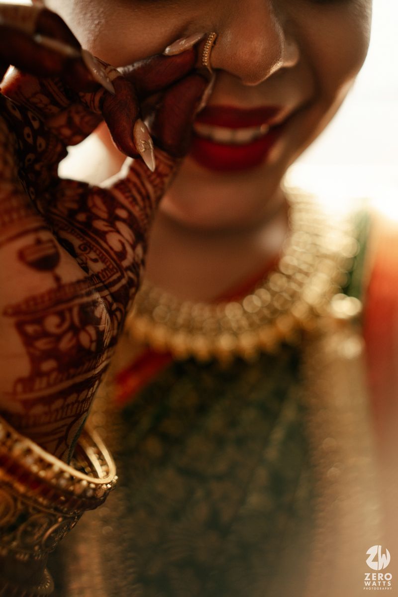 Indian bride smiling with intricate mehndi henna hands, gold jewelry and red lips in warm golden light.