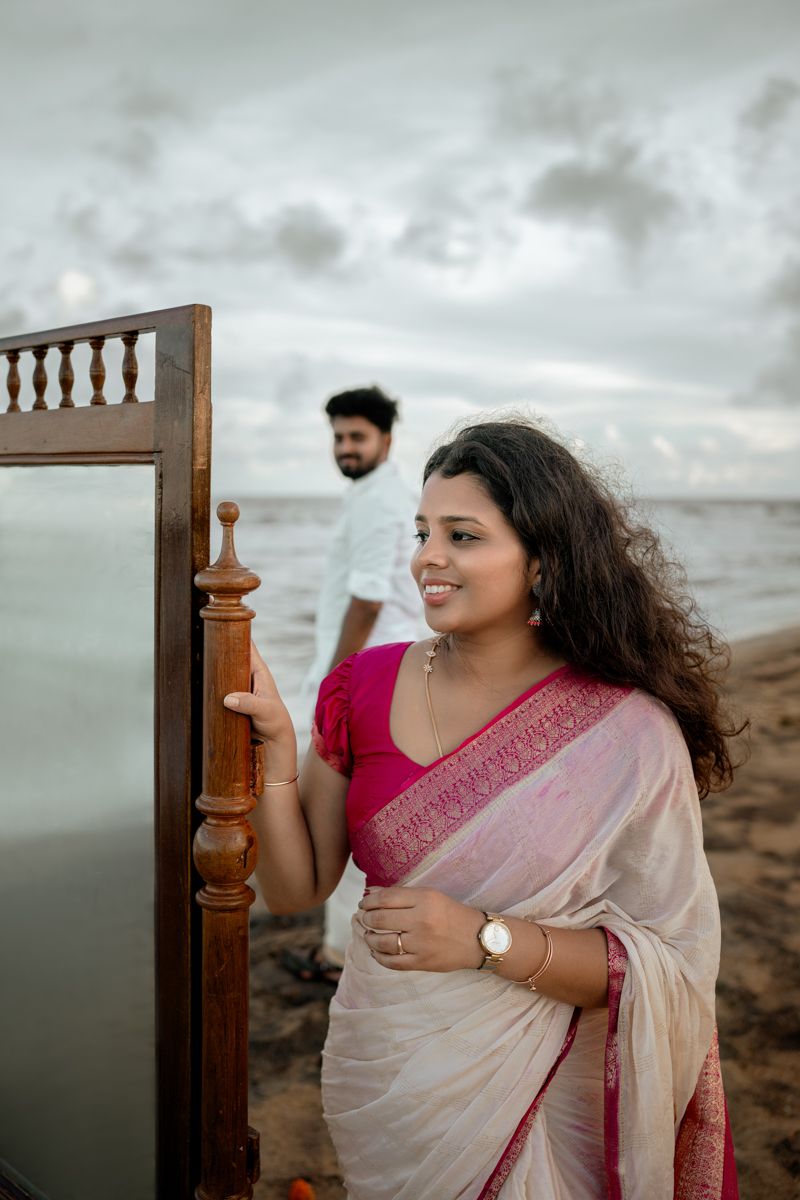 A person in a white and pink saree poses against a cloudy sky at a beach with wooden railing.