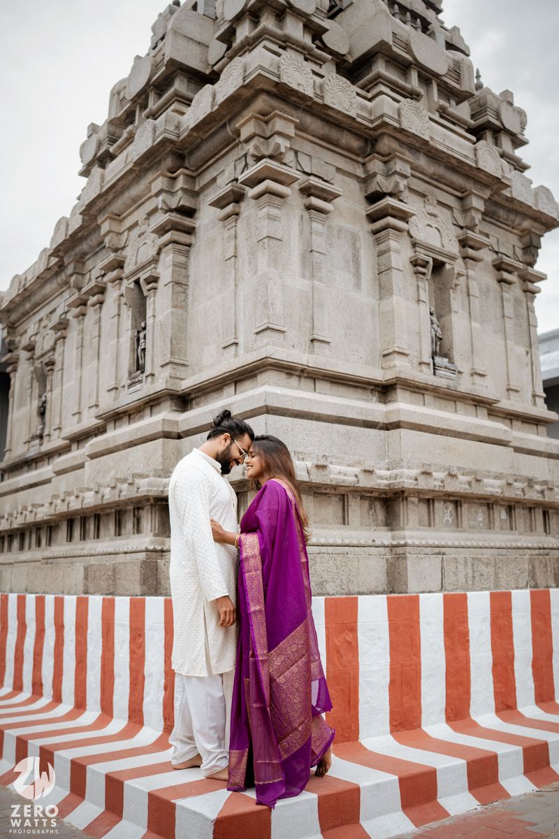 Couple in traditional Indian attire poses against temple walls with red and white striped barriers.