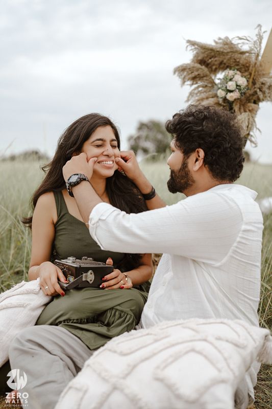 A couple shares an intimate moment outdoors while sitting on blankets in a grassy field with pampas grass decor.