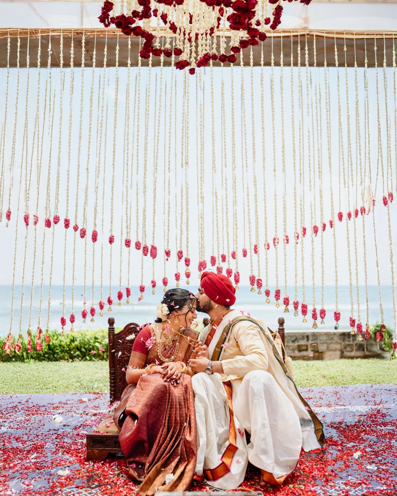 A couple sits together on grass surrounded by scattered red rose petals in a romantic outdoor setting.