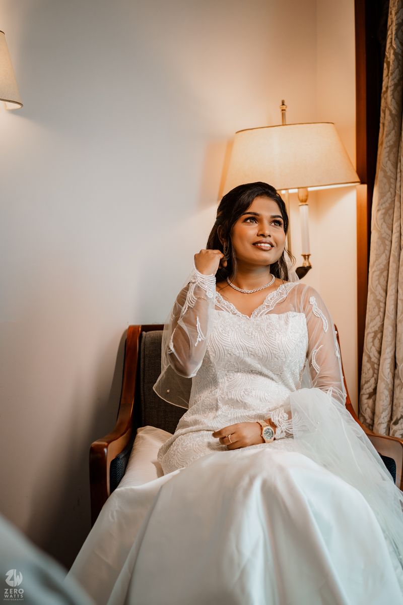 A bride in a white lace wedding gown sits elegantly in a room with warm lighting and a table lamp.