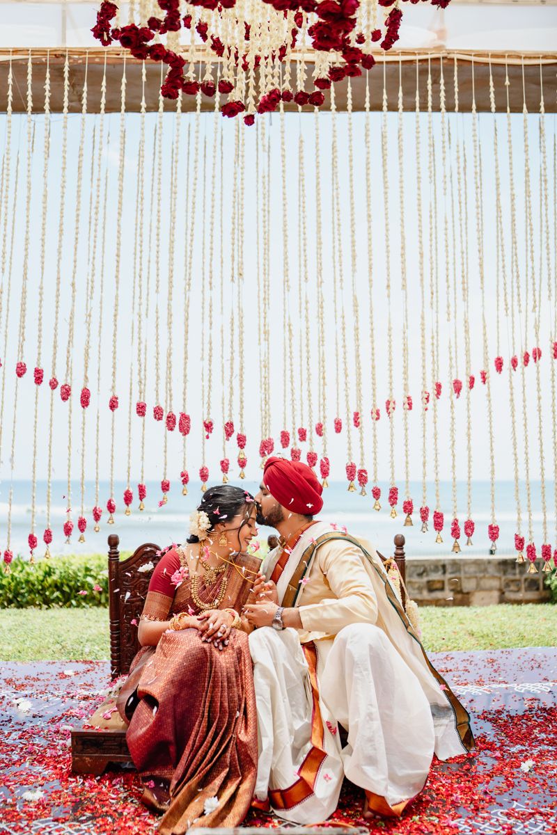 Traditional Indian wedding couple shares intimate moment under red floral mandap decorations by the ocean.