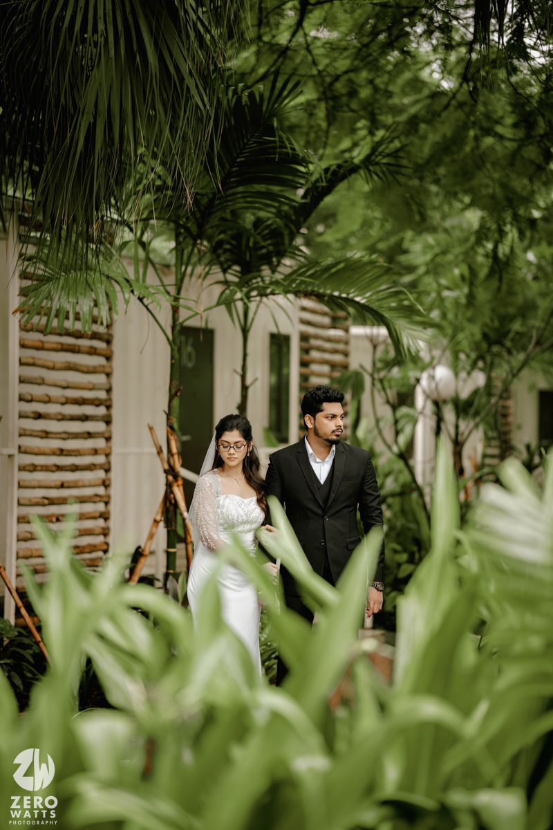 A couple in wedding attire walks together in a lush tropical garden setting with palm fronds and greenery.