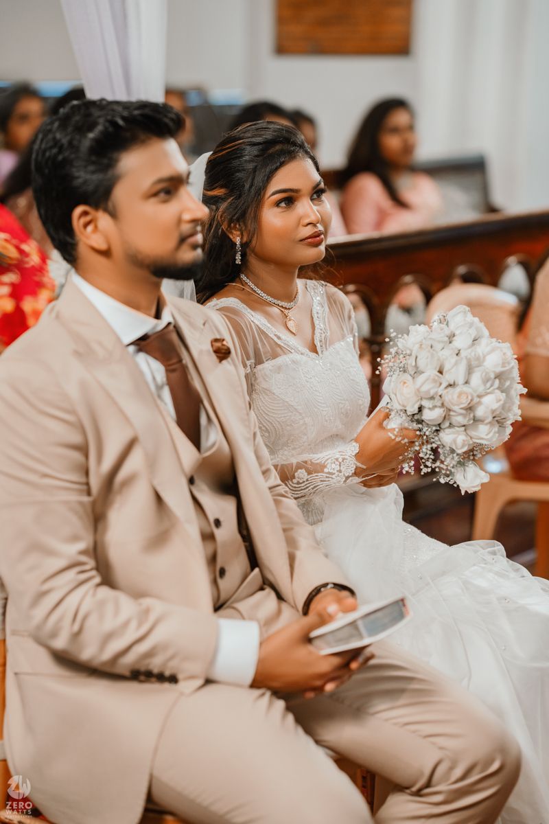 Wedding couple seated in church pews wearing coordinated beige and white attire during their ceremony.