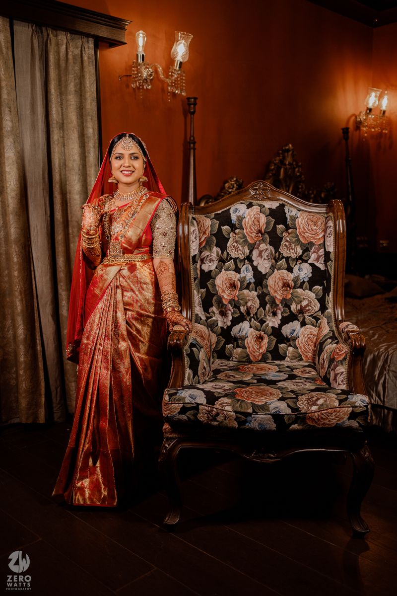 A person in a coral-colored dress poses against an ornate sofa in a warmly lit room.