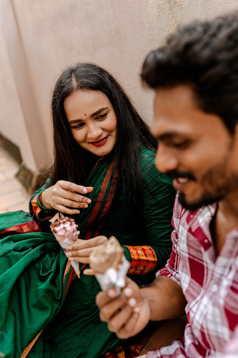 A couple shares a playful moment while feeding each other sweets in a casual outdoor setting.