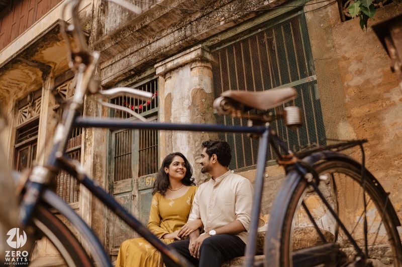 A couple shares a romantic moment in an urban alleyway with vintage buildings and a bicycle in the background.