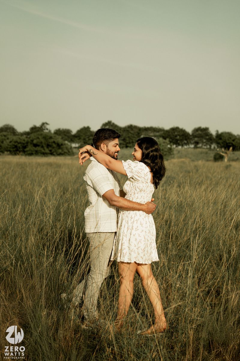 A couple embraces in a sunlit meadow wearing coordinated white outfits for their engagement photos.
