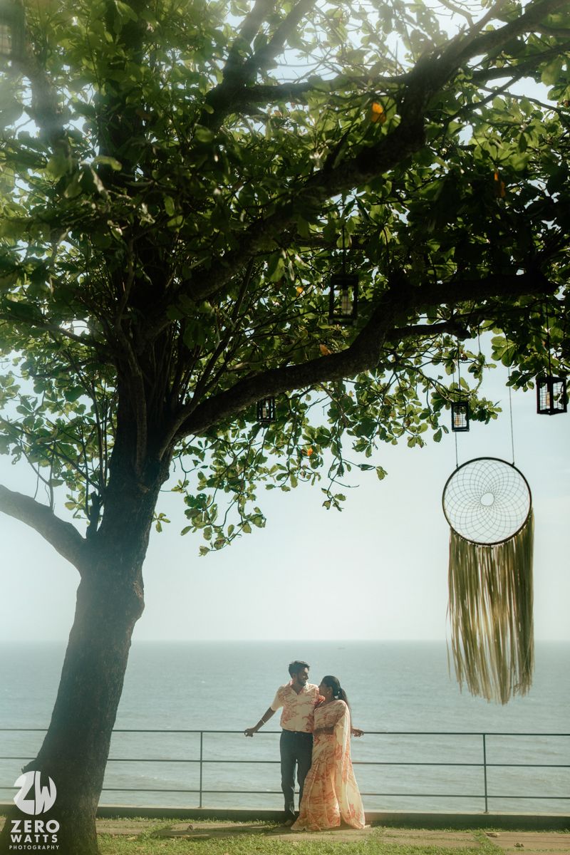 A scenic outdoor setting with a large tree providing shade over a seaside railing and decorative lanterns.