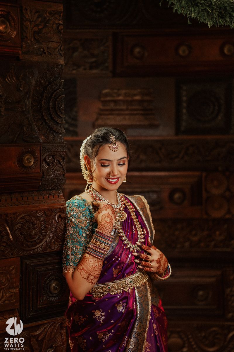 South Indian bride in purple silk saree with teal blouse adorned in gold jewelry and bridal makeup.