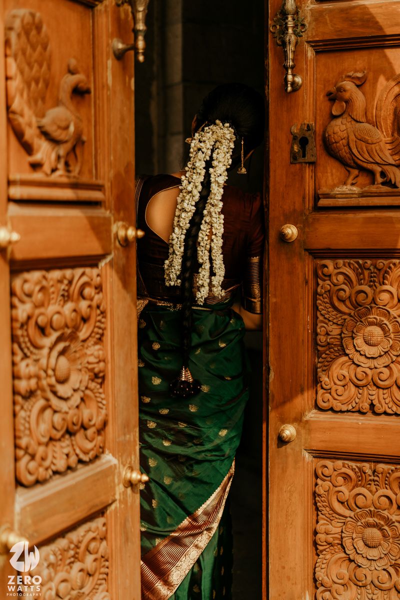 Bride in green silk saree with jasmine flowers in hair peeks through ornate carved wooden temple doors.