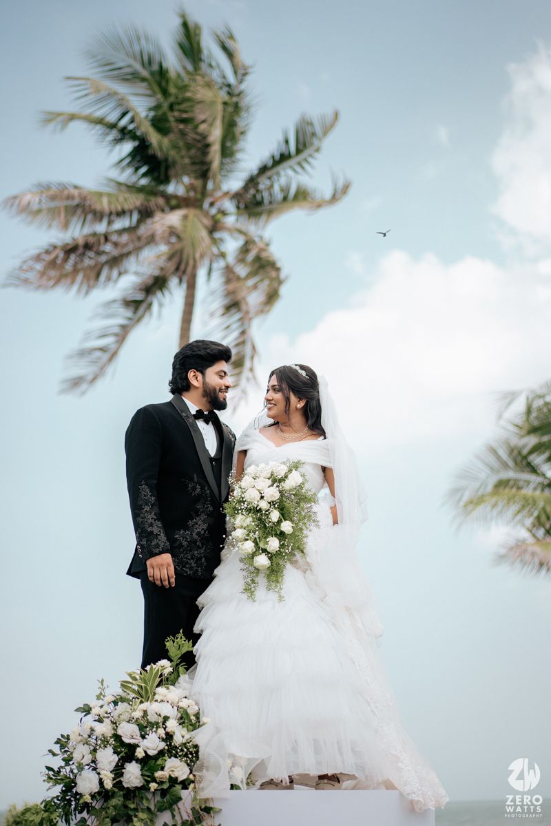Bride in white ruffled gown and groom in black tuxedo pose romantically under palm trees at a beach wedding.