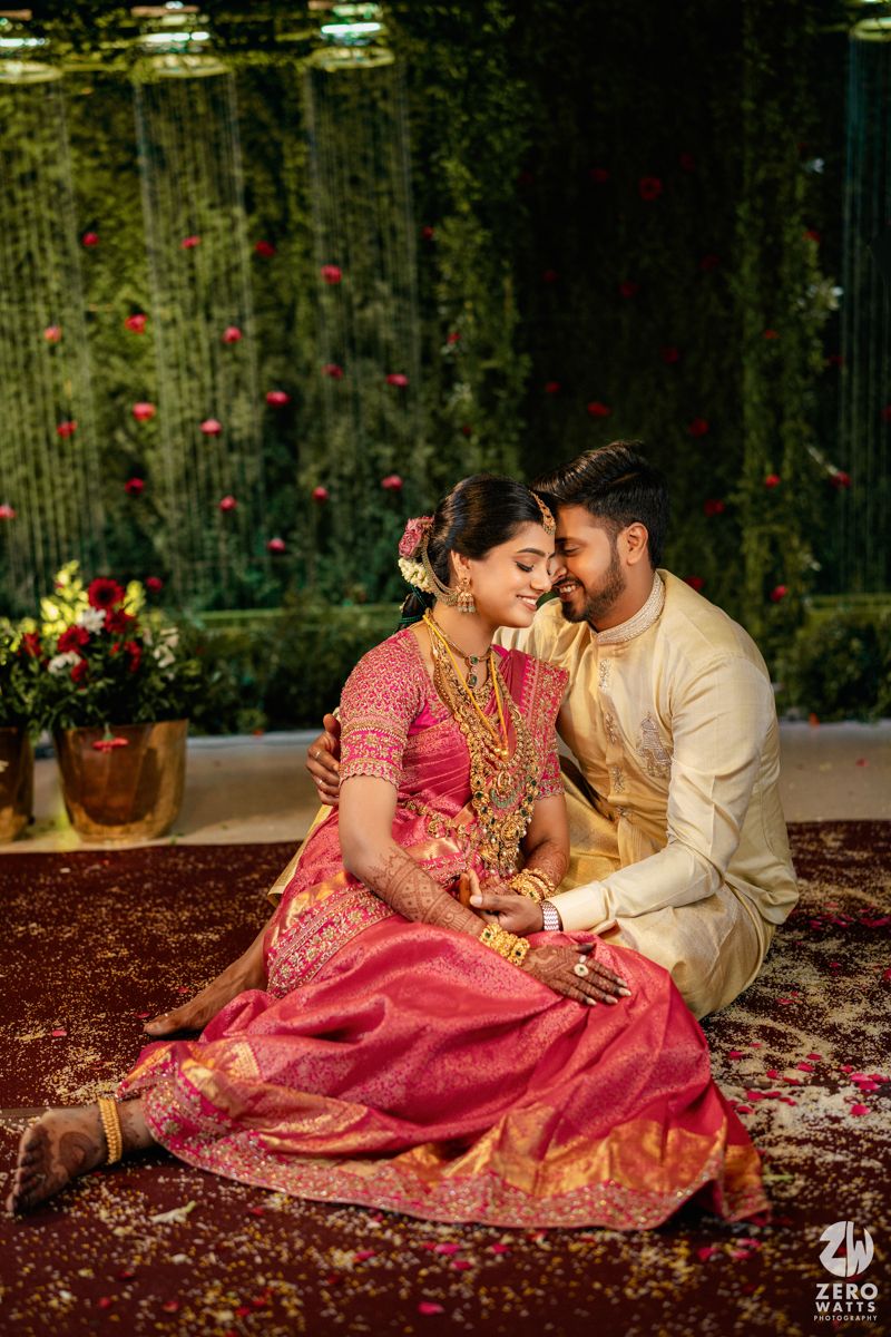 A bride in red silk saree and groom in cream sherwani share an intimate moment at their traditional South Indian wedding.