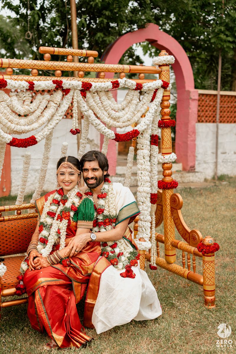 Indian bride and groom in traditional attire seated under floral mandap during South Indian wedding ceremony.