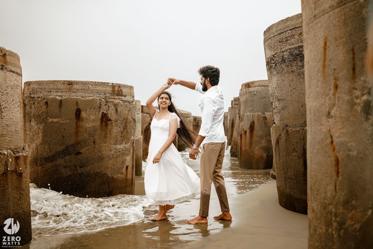 Couple dancing between rustic stone pillars on a beach, woman in white dress being twirled by man in brown outfit.