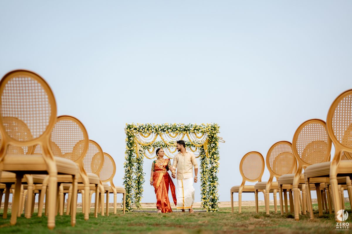 South Indian bride and groom standing under floral arch at outdoor wedding ceremony with elegant rattan chairs.