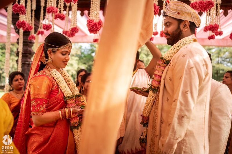 Bride in orange saree and groom in white sherwani exchange garlands at a traditional South Indian wedding ceremony.