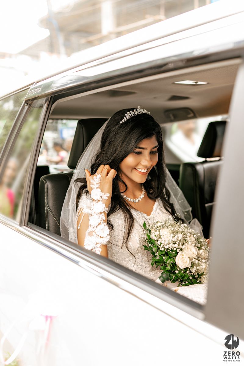 Smiling bride in white gown and tiara holding bouquet inside a wedding car.