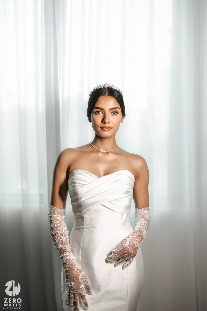 Elegant bride in strapless white gown with lace gloves and tiara poses before sheer curtains.