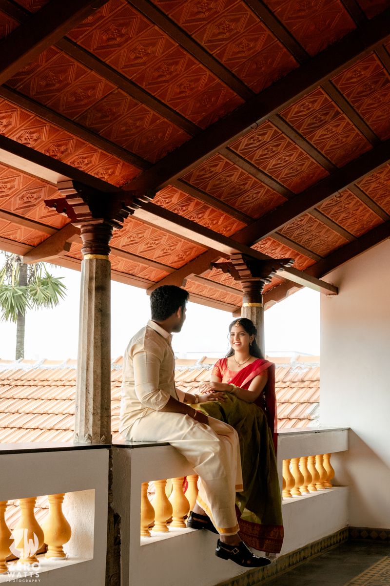 Couple in traditional Indian outfits sharing a moment on a balcony with ornate red-tiled ceiling and pillars.