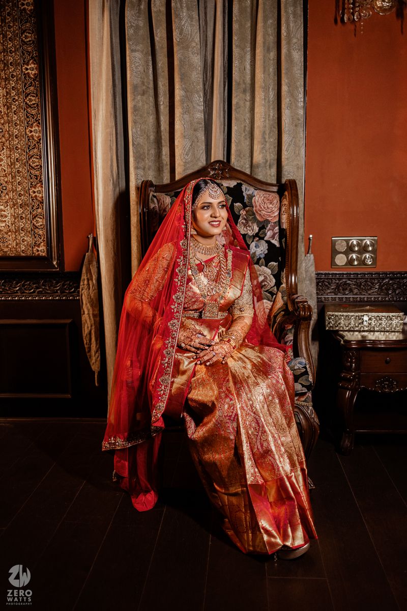 A traditional Indian bride in a red and gold wedding sari poses gracefully in an ornate interior setting.
