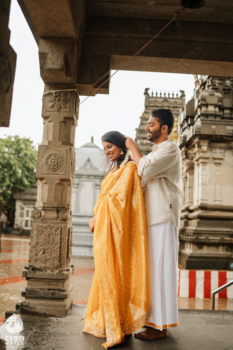 A couple in traditional Indian attire share a romantic moment under ornate stone pillars at a historic temple.