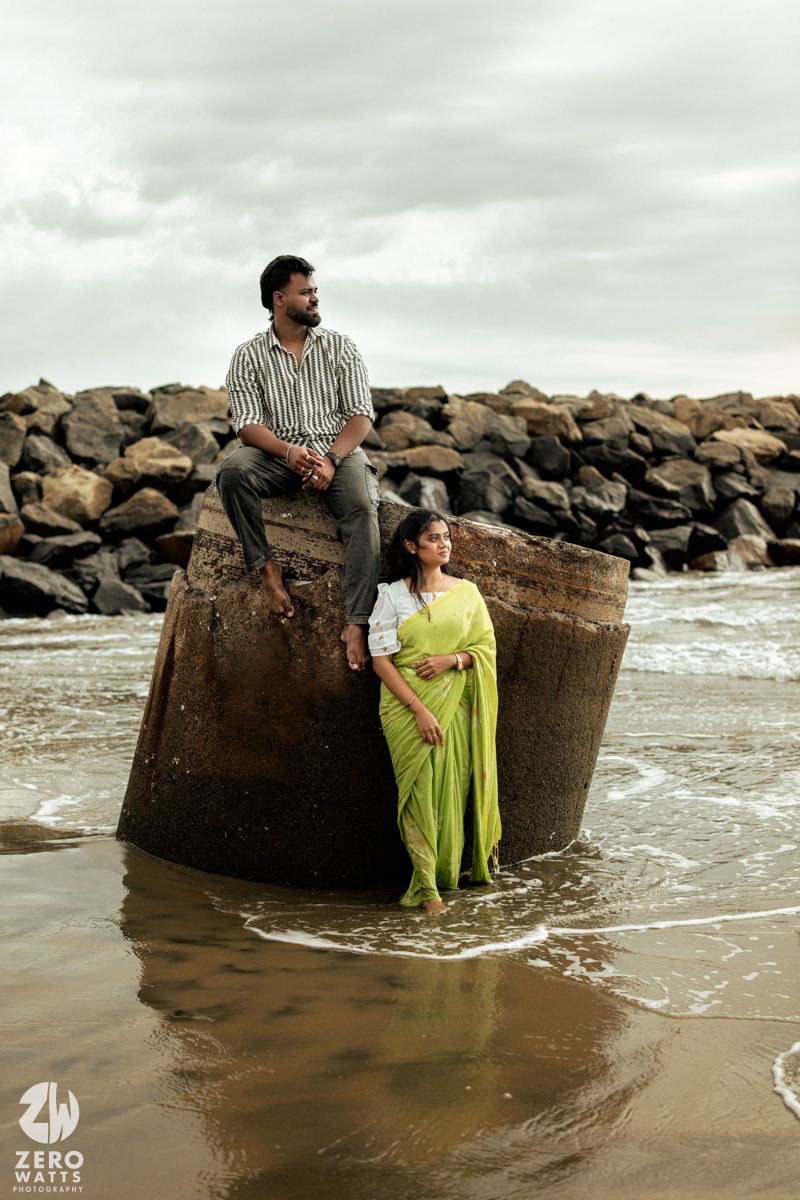 Couple sits on concrete pillars surrounded by rocks and waves during a beach photoshoot.