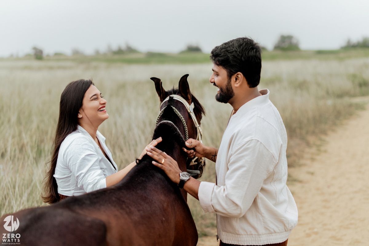 A romantic couple embraces in a grassy field with horses grazing in the background on an overcast day.