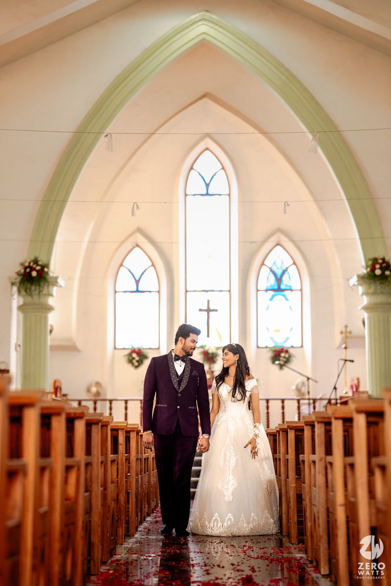 Newlywed couple holds hands walking down sunlit church aisle with arched windows and wooden pews.
