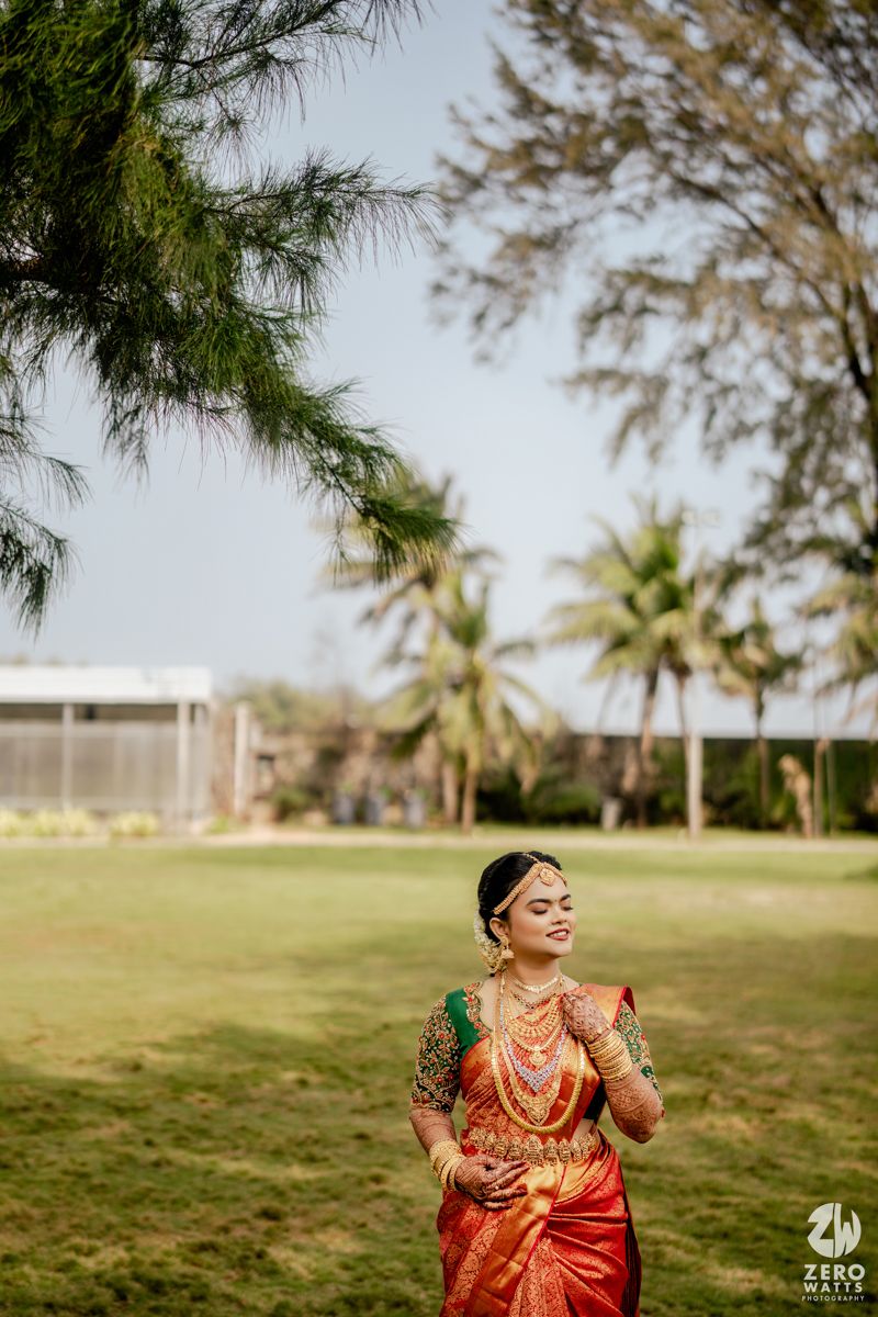 South Indian bride in red and green silk saree with gold jewelry poses outdoors near palm trees.