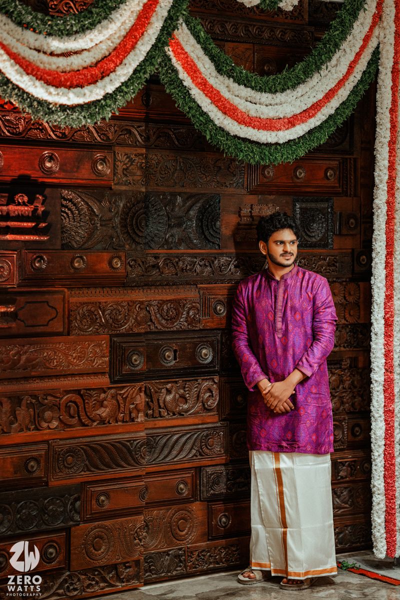 Man in purple kurta and white dhoti stands before ornate carved wooden door adorned with tricolor floral garlands.
