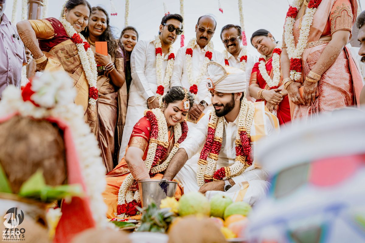 Traditional South Indian wedding couple in red and white attire performing sacred rituals surrounded by family members.
