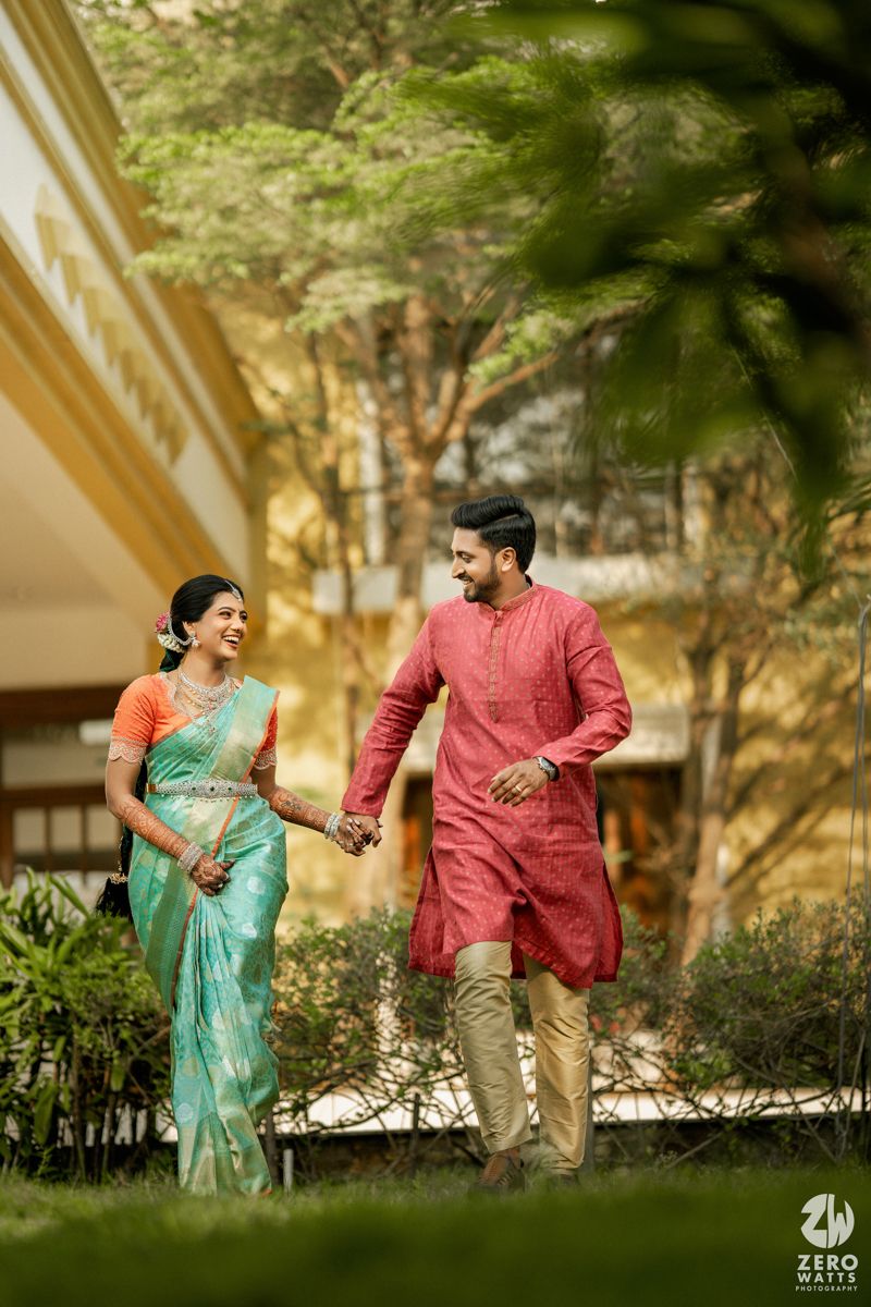 Indian couple in traditional attire walking hand-in-hand outdoors, bride in teal saree and groom in red kurta.