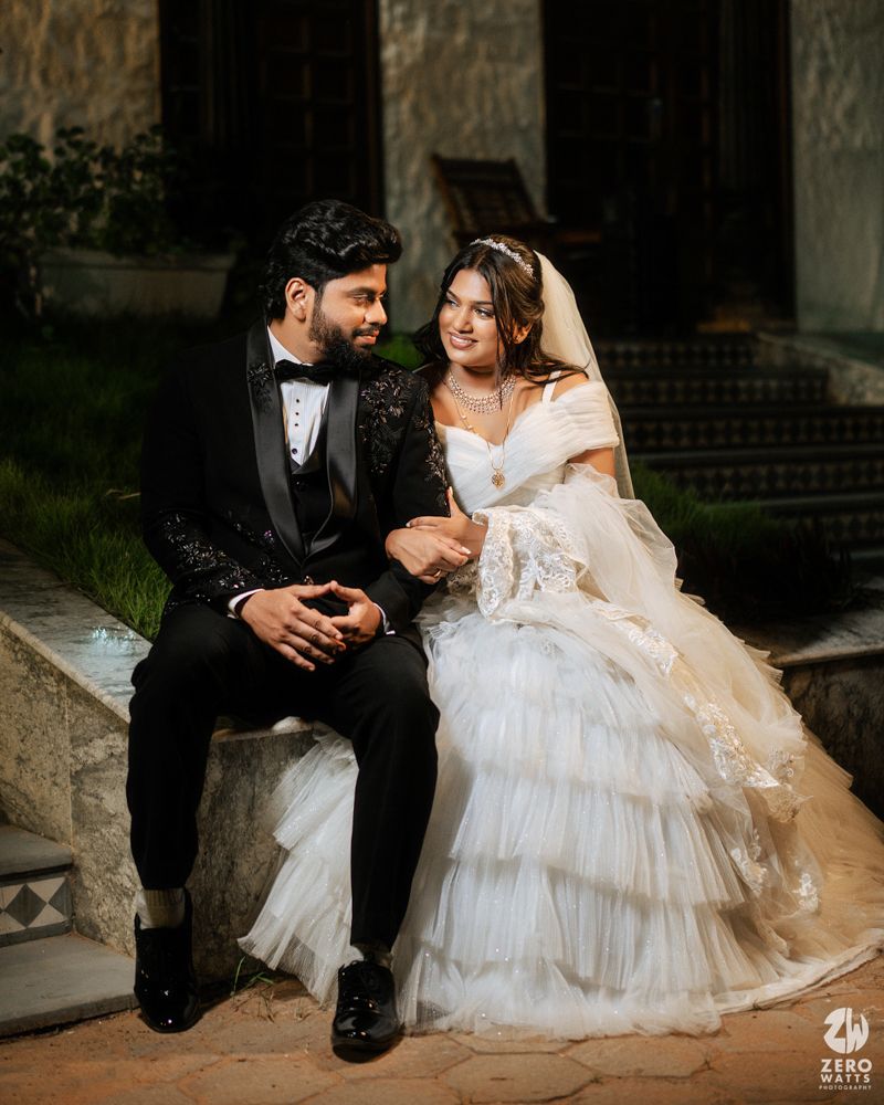 A couple in wedding attire sits together, groom in black tuxedo and bride in white ruffled gown with veil.