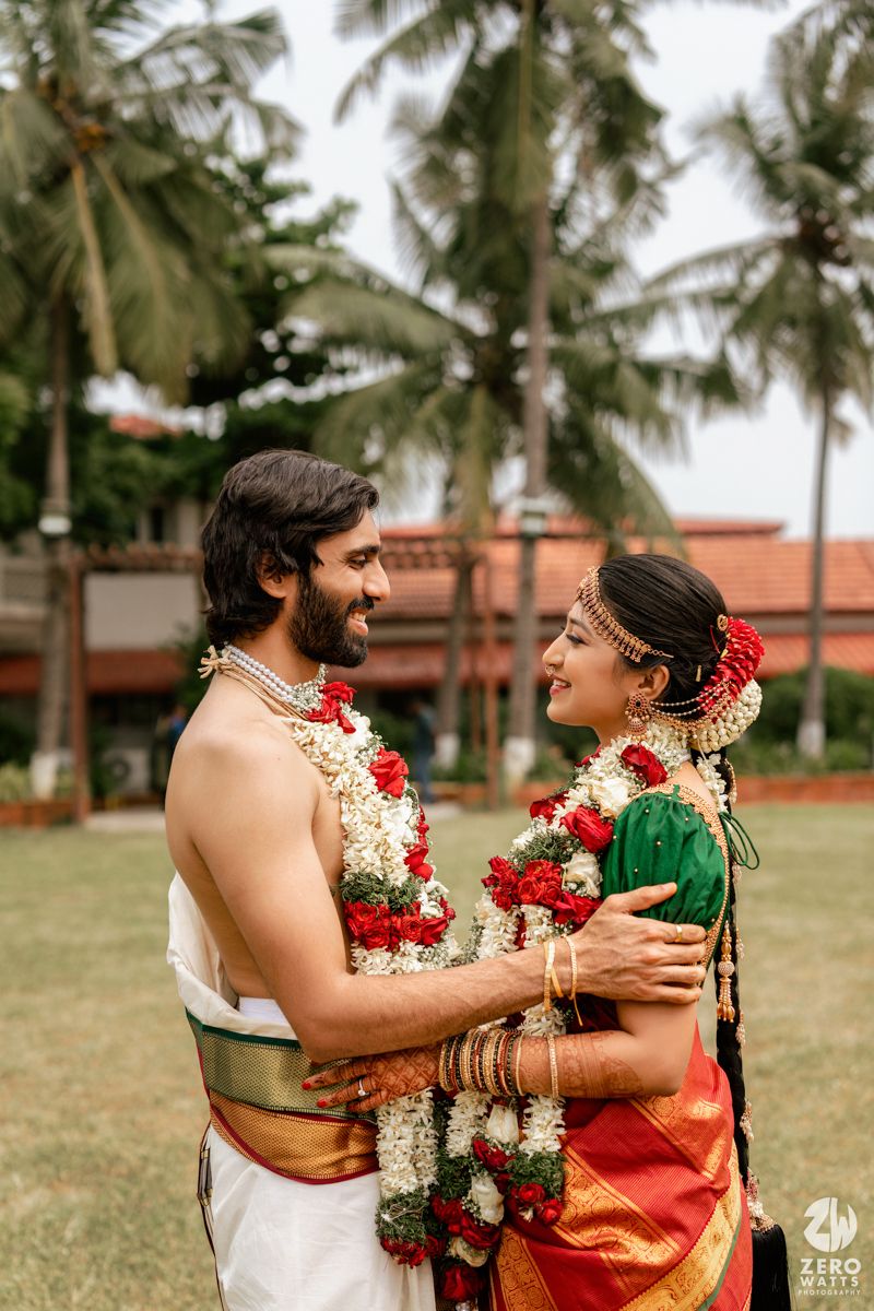 South Indian bride and groom in traditional attire exchanging garlands outdoors with palm trees in background.