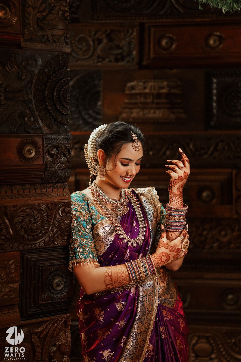 Bride in purple silk saree with teal blouse showcases mehndi hands, wearing traditional gold and pearl jewelry.