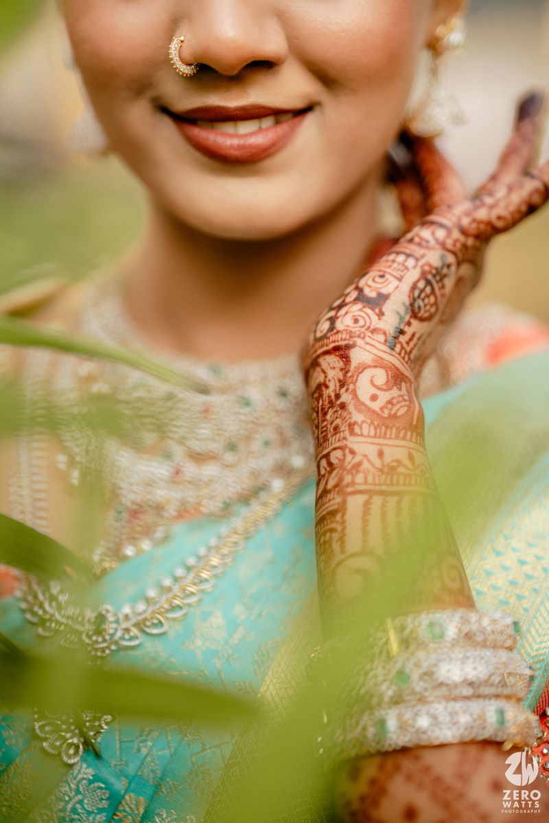 Close-up of Indian bride showcasing intricate bridal mehndi design on hands with teal saree and nose ring.