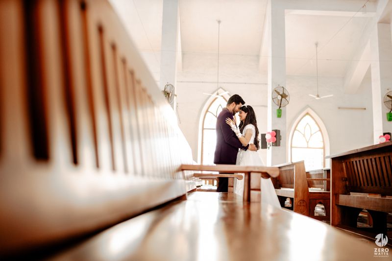 Bride and groom share intimate moment among wooden church pews with arched windows in background.