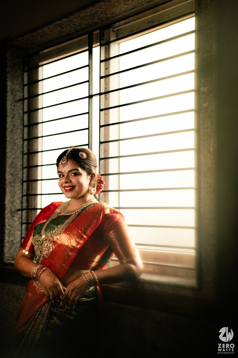 Radiant South Indian bride in red and green silk saree with gold jewelry posing by window in soft backlight.