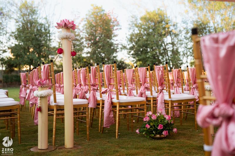 Elegant outdoor wedding ceremony setup with gold chiavari chairs adorned with pink ribbon sashes on a lush green lawn.