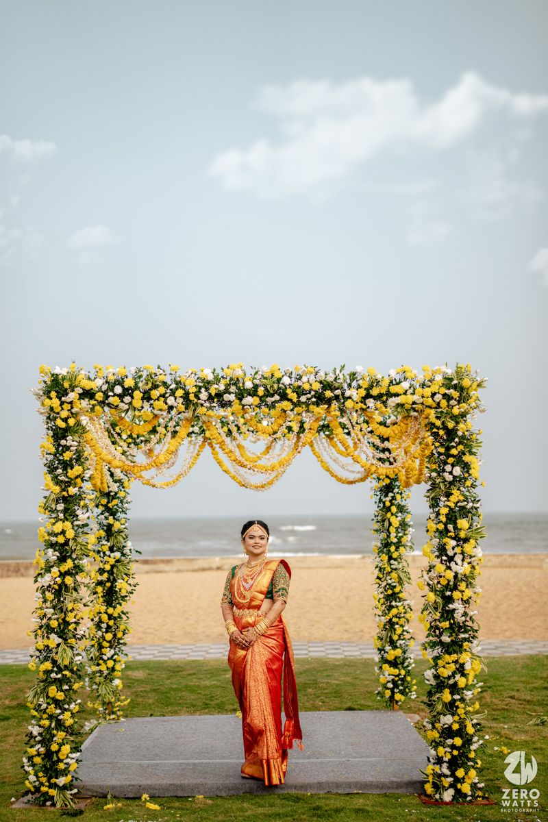South Indian bride in red silk saree stands under a floral mandap arch decorated with yellow flowers by the beach.