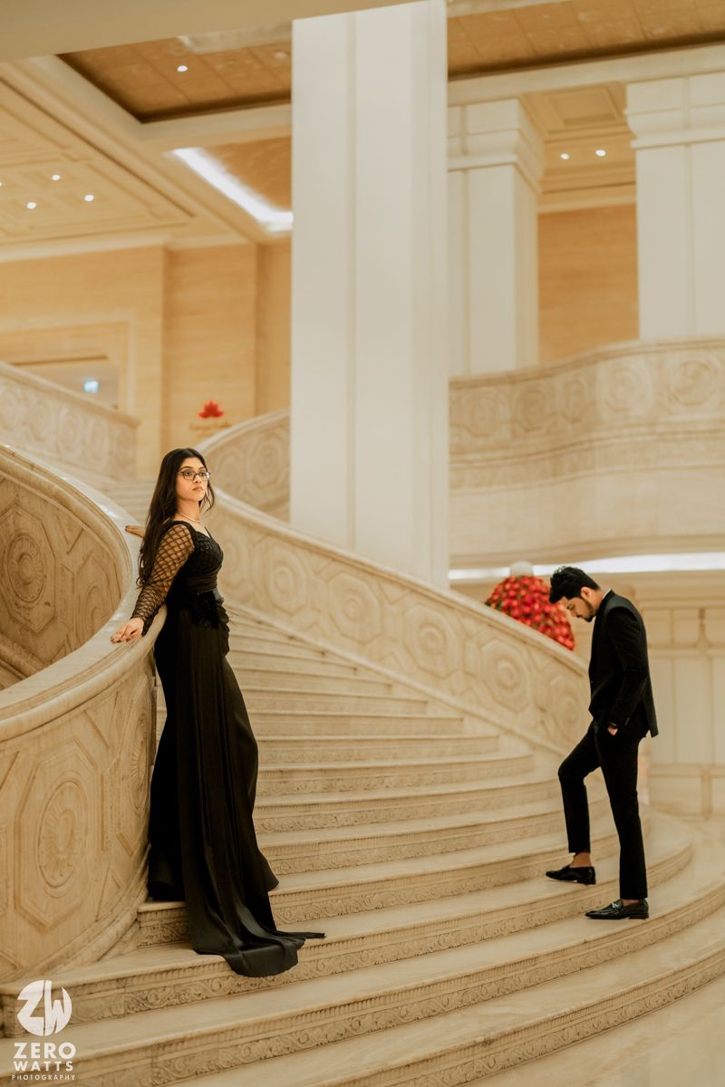 Couple in black attire poses on grand marble staircase in elegant indoor setting.