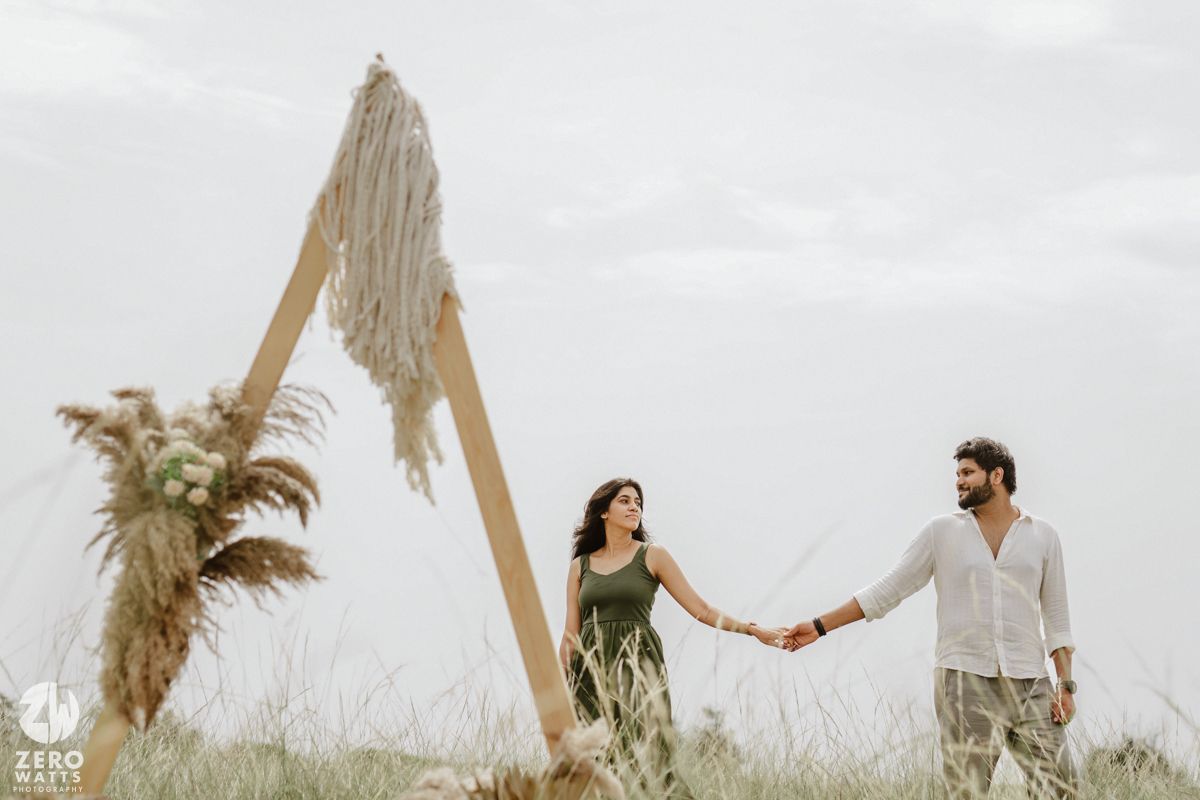 Couple holds hands in a sunlit field surrounded by tall wooden posts and natural decor.