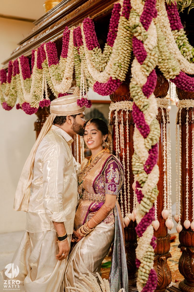 Couple in traditional Indian wedding attire standing beneath ornate floral mandap with purple and white decorations.