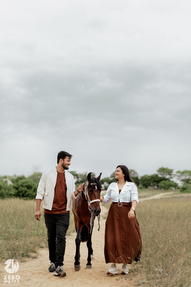 A couple walks together with their horse on a grassy path under cloudy skies in rustic countryside attire.
