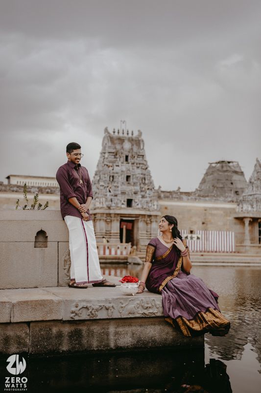 A couple in traditional Indian attire sits by a temple pond with ornate architecture in the background.