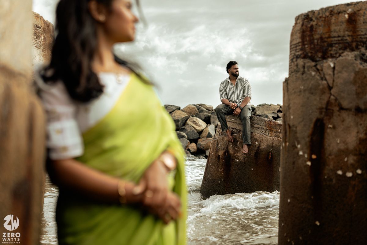 A creative beach photoshoot with one person sitting on a concrete structure while another poses below.