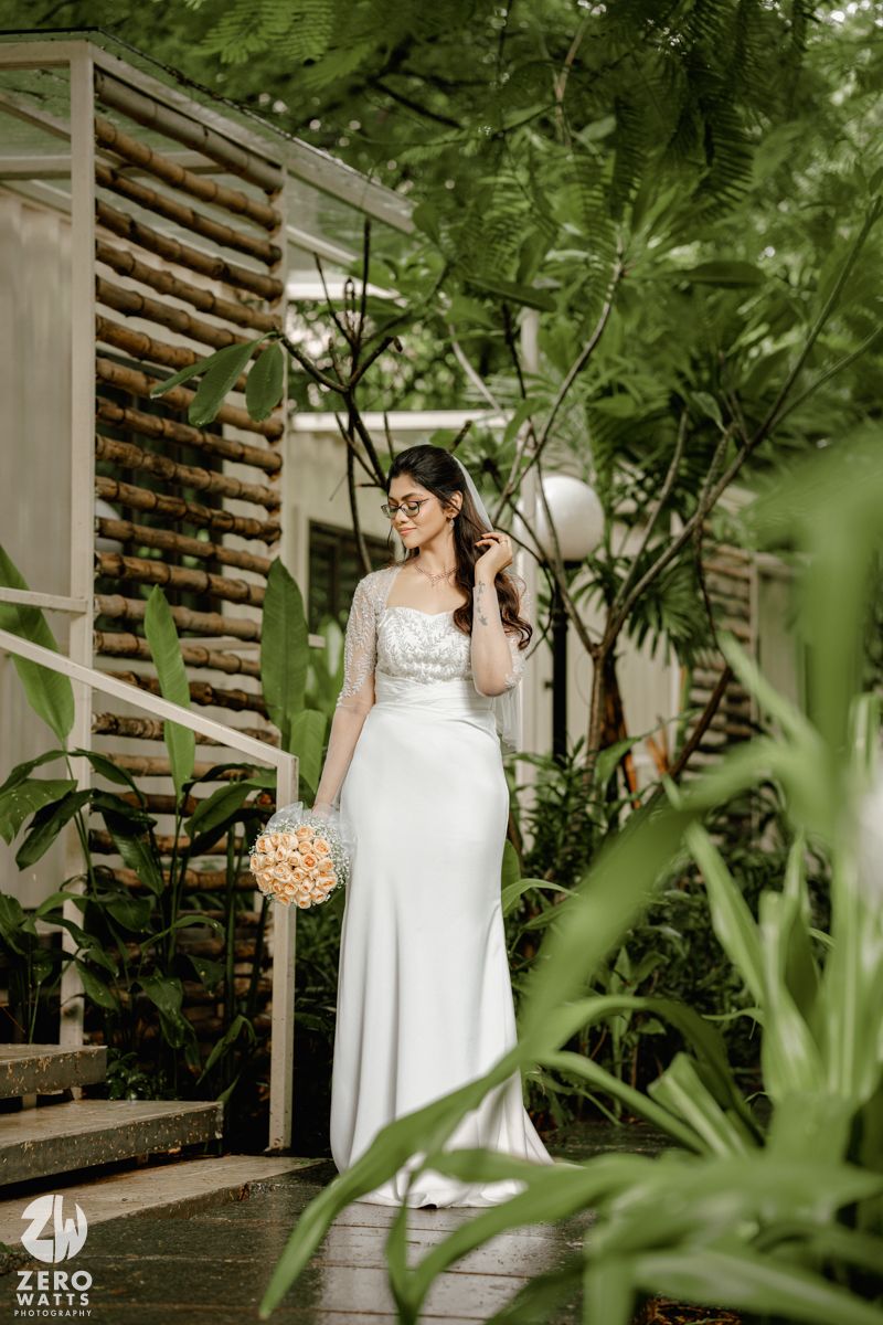 A bride in a white wedding dress stands in a lush garden setting with wooden slatted walls creating an elegant backdrop.