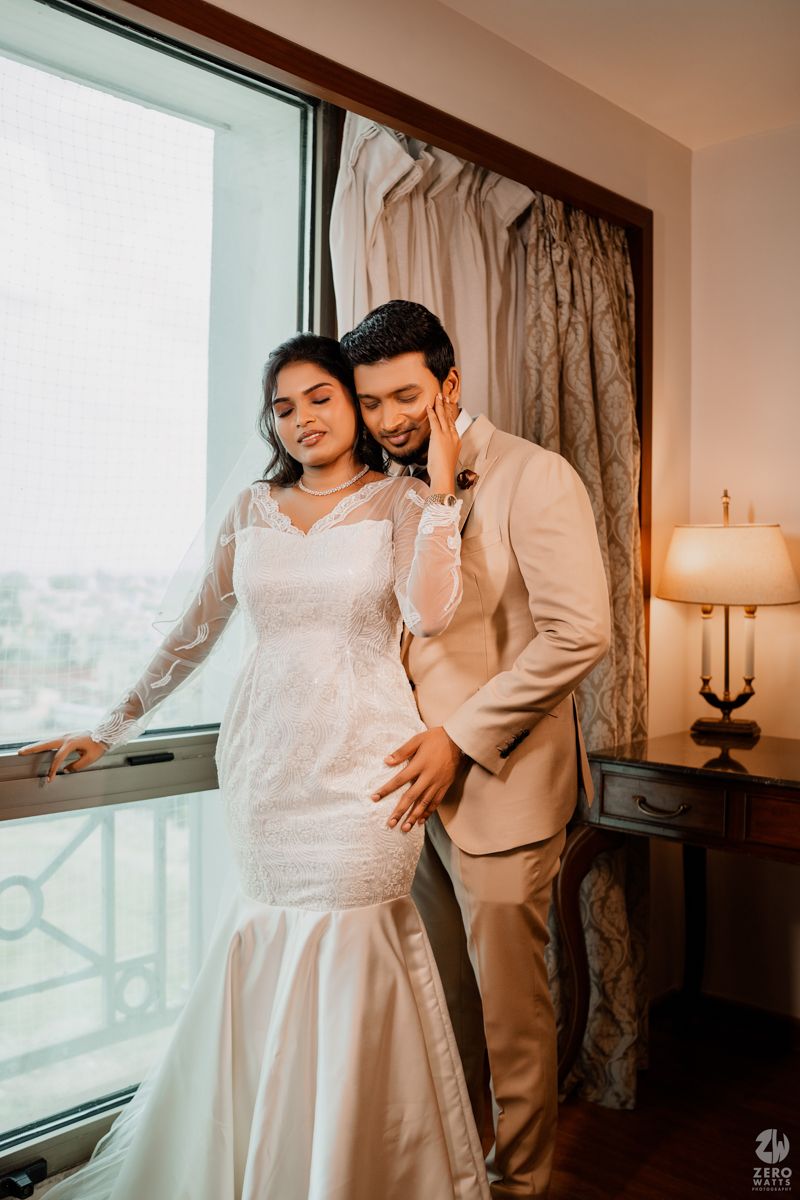 A romantic wedding portrait of a couple in formal attire posing by a window in warm lighting.